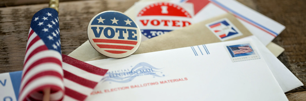 Desk with small American flag, two "Vote" pins, and an envelope with "Official Election Mail" printed on it.